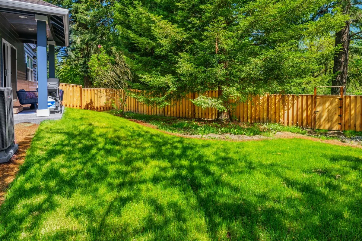 A lush, securely fenced backyard with green grass, tall trees, and a cozy patio area, offering ample space for pets and children to play safely outdoors. The area is bright and inviting, designed for both relaxation and active fun.