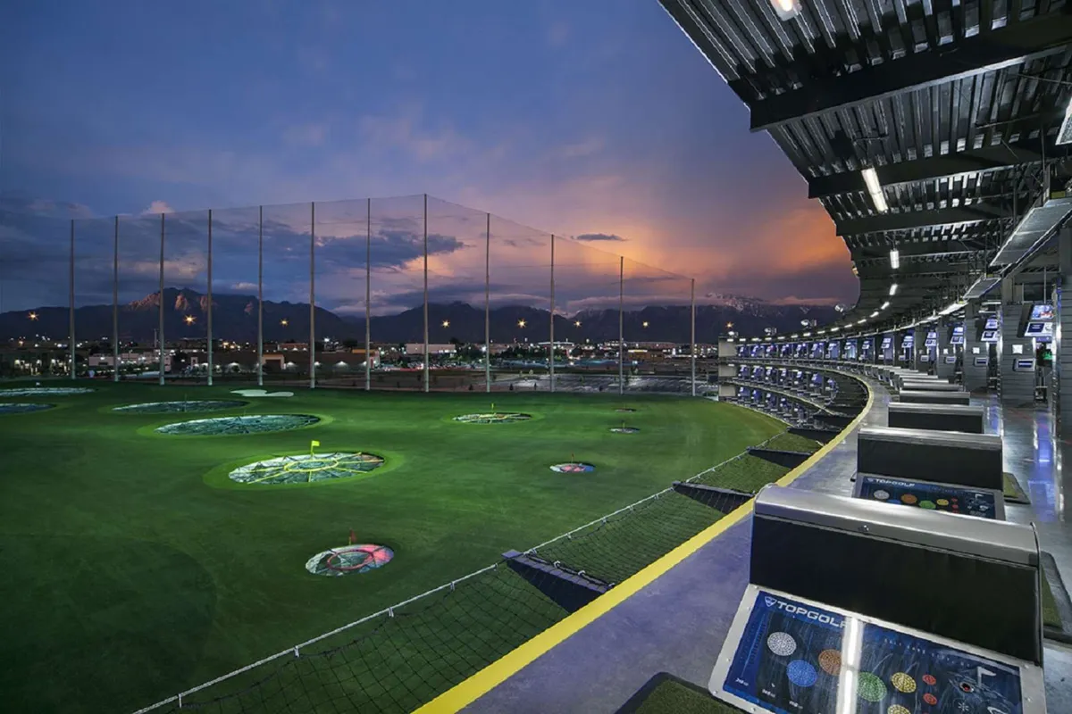 An inviting exterior view of a golf driving range with illuminated hitting bays against a dramatic mountain backdrop. While not part of the backyard, the image captures the spirit of outdoor recreation and socializing in a beautiful natural setting.