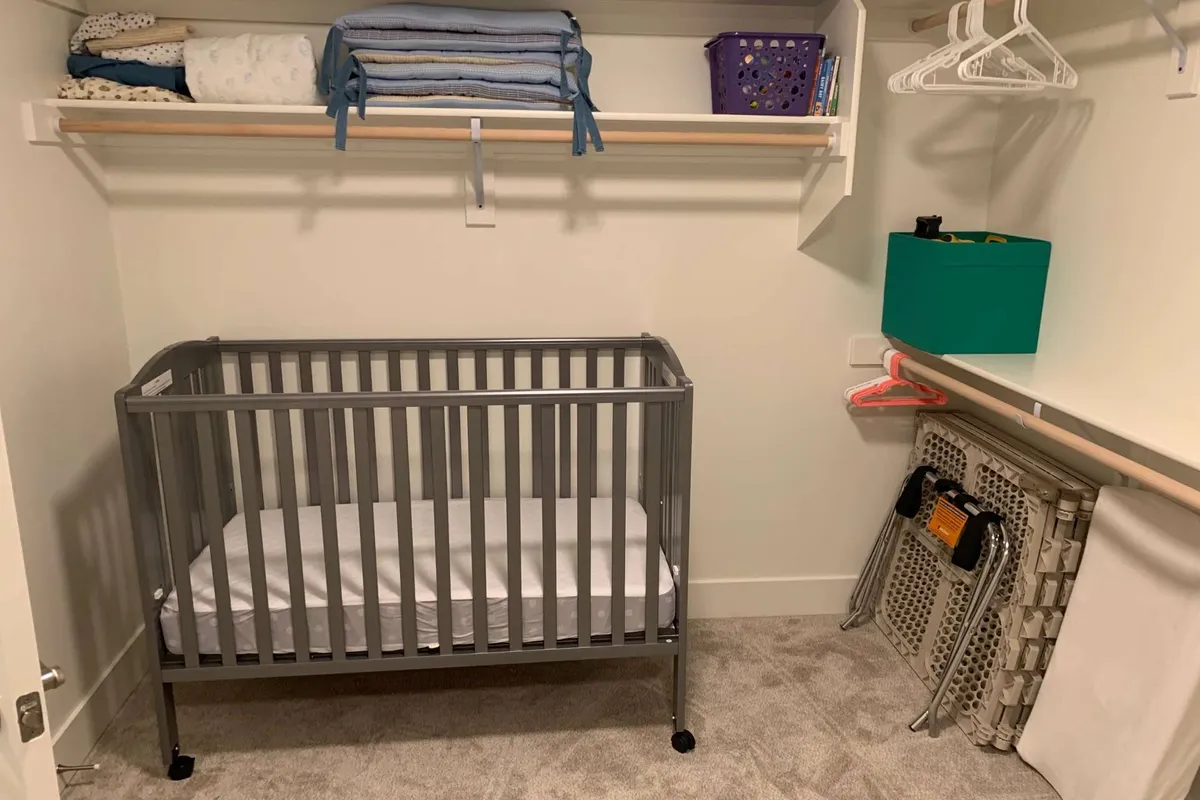 A charming, well-lit nursery with a crib, shelves stocked with blankets, and a storage box for toys. Hanging clothes and soft colors create a welcoming space for families with young children.