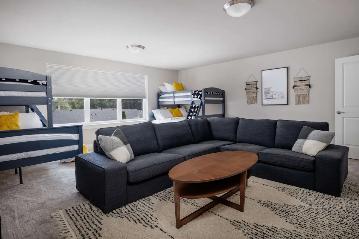 A casual living area anchored by a large sectional sofa, bunk beds, a wooden coffee table, and decorative wall art. The scene suggests comfort, movie nights, and board game gatherings in a family-friendly environment.