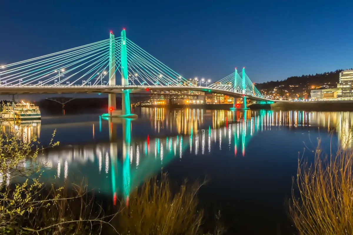 A striking view of a city bridge over water at night, with twinkling city lights and tranquil reflections. The scene suggests urban exploration and the allure of nearby Portland, capturing the excitement of local discovery.