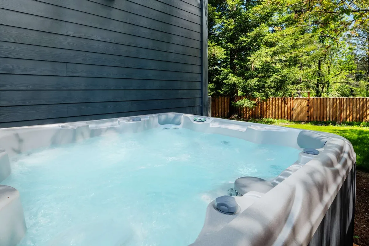 A serene backyard scene featuring a bubbling hot tub framed by lush greenery and a wooden privacy fence. The setting is peaceful, inviting, and clearly designed for relaxation and socializing in nature.