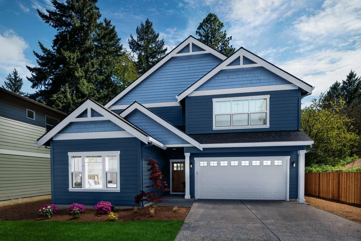 The image showcases the handsome blue exterior of a newly built single-family house with crisp white trim, a spacious garage, and a welcoming front yard bursting with colorful flowers. The inviting porch promises stunning sunset views and a perfect spot for morning coffee, all set in a tranquil, suburban neighborhood just outside Portland.